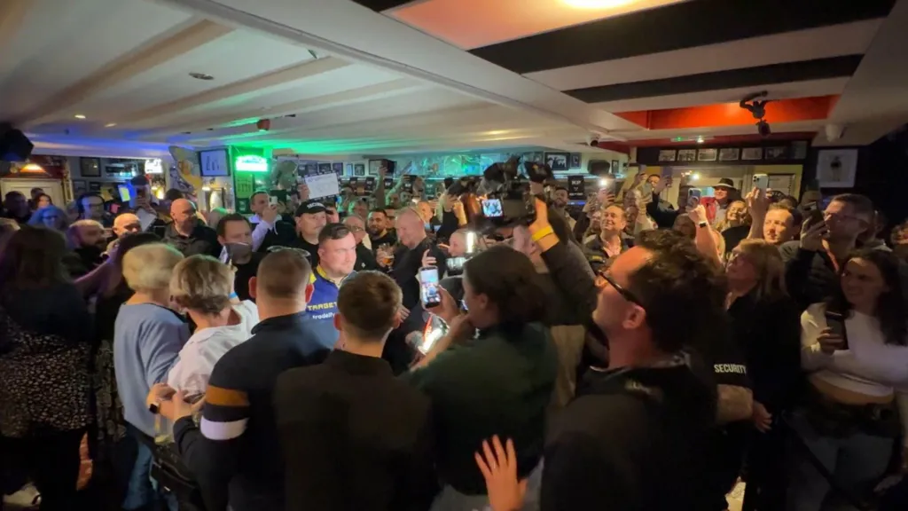 A crowd of people in a pub gather around Luke Littler. Some of the people are taking pictures of him.