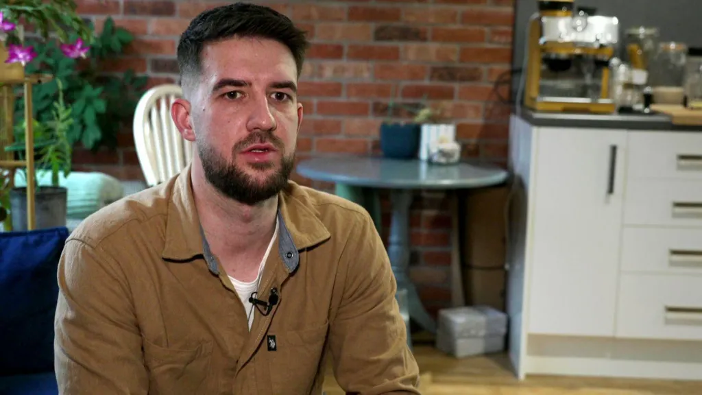 Bart wears a mustard shirt and is sitting in a kitchen and dining room space, with a red brick wall behind him.