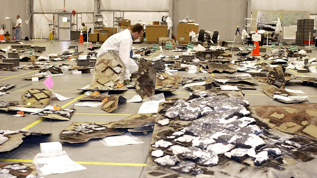 Debris from the Space Shuttle Columbia is laid out on the floor of a large hanger. A man in a white coat is bending down inspecting some of it.  There are tiles and pieces of white foam - some of the material is charred. 