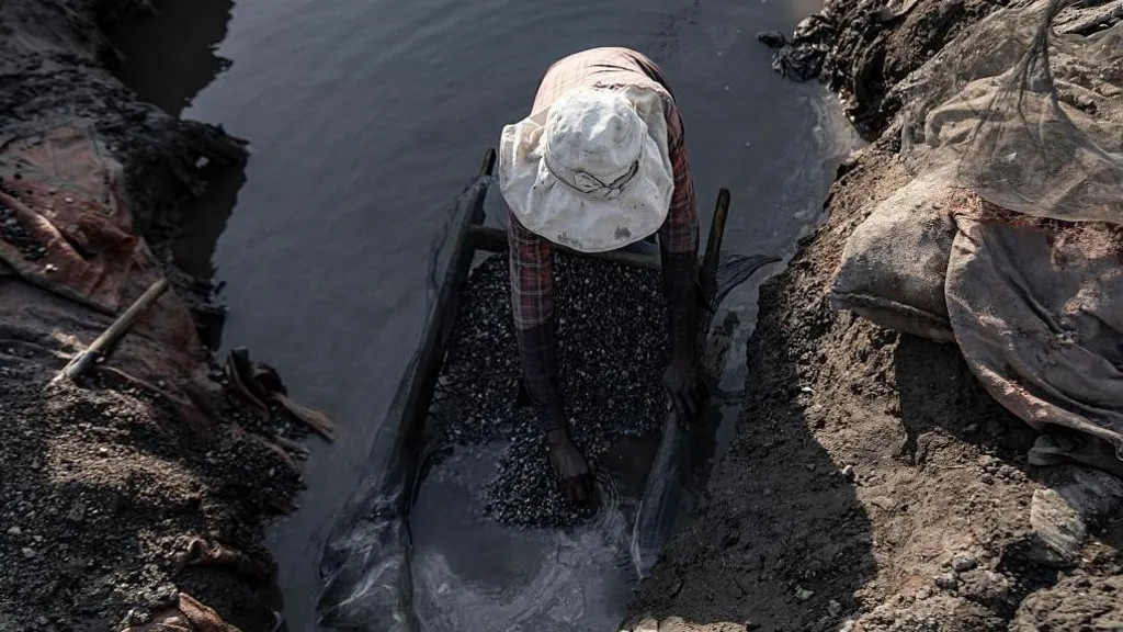 An aerial view of a  woman in a white hat in a small pond of water sorting through some stones that are on a tray. 