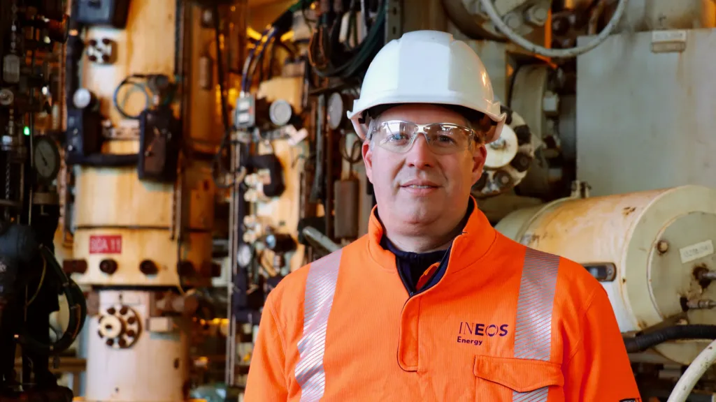 Mads Gade, CEO of INEOS Energy, wears a bright orange, high-viz jacket, safety glasses and a white helmet. He stands in front of pipes and pumping equipment.
