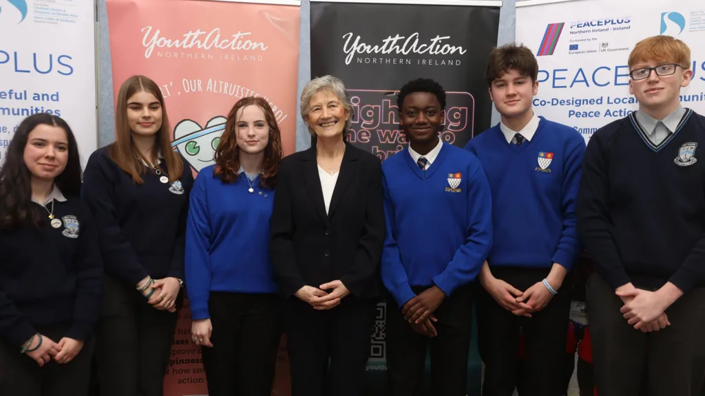 Shows seven people, including six students in school uniforms - three girls to the left and three boys to the right with a woman in the middle dressed in a blac suit with white top.