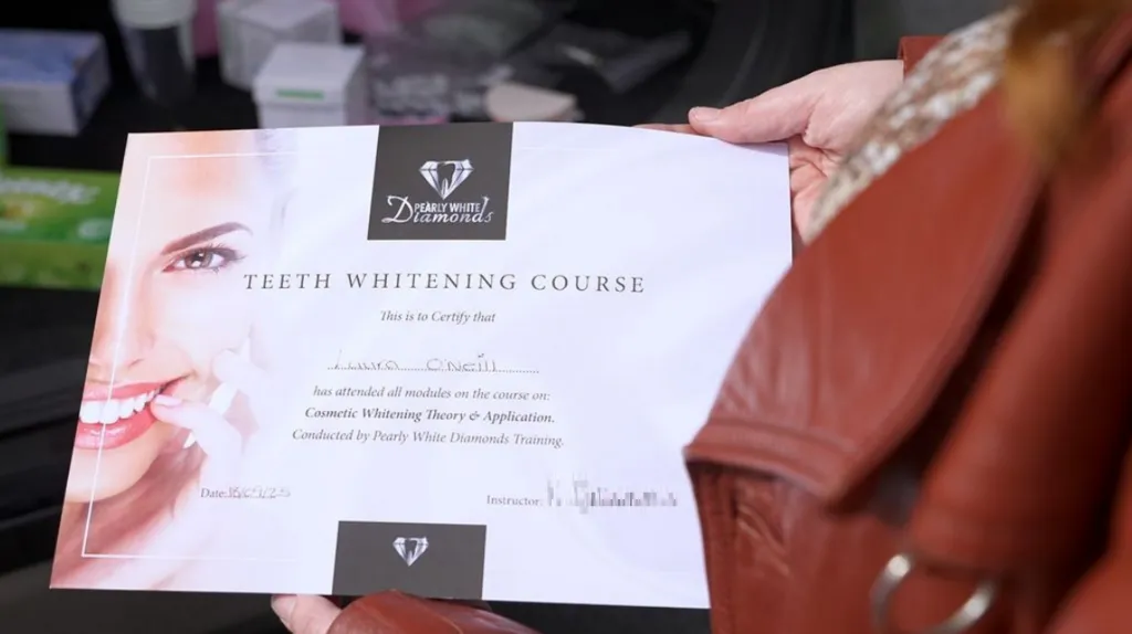 A certificate from Pearly White Diamonds, being held by our undercover reporter. On the left-hand side is the right-hand side of a woman's face, bearing a big grin, and on the remainder of the certificate the following is written: "Teeth Whitening Course. This is to certify that Laura O'Neill has attended all modules on the course on Cosmetic Whitening Theory and Application. Conducted by Pearly White Diamonds Training. Date 15/09/2025." The instructor's signature, which is pixelated in the image, is on the right-hand side.  
