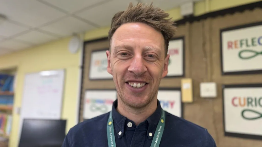 Man with spikey brown hair smiling at the camera. He is in a classroom and is wearing a lanyard around his neck. 