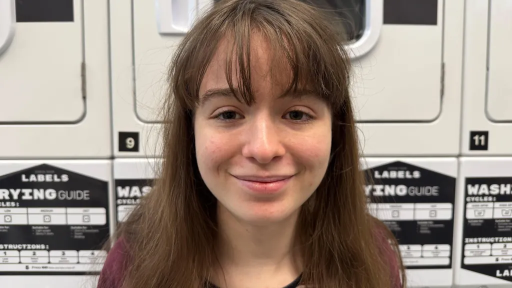Ellie May is a first-year history student. In this image she is smiling in front of the washing machines in a laundry room on campus at the University of York. Ellie May is wearing a stripey red top and has straight brown hair with a fringe. 