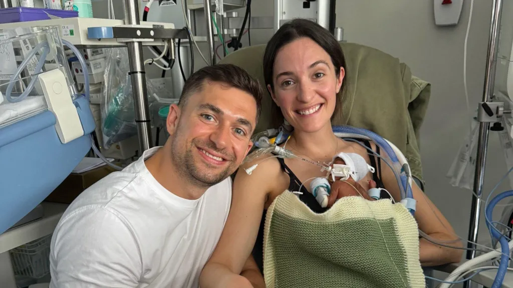 A dark haired young man sits in hospital with a dark haired young woman. The woman hold a small baby on her chest. The baby is wired to medical equipment.