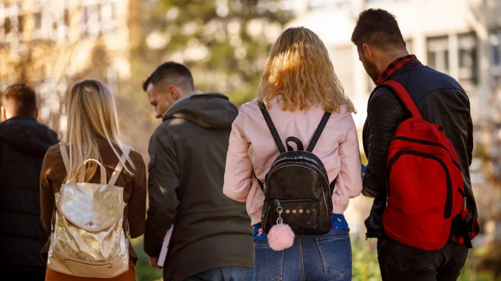 A stock image shows a group of young people wearing backpacks walking together along a tree-lined path on campus, near university buildings