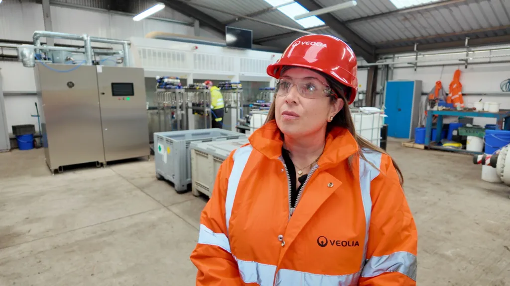 Nicola Henshaw is standing in a room where nitrous oxide canisters are being treated.  She is wearing a high-vis jacket, with a range of machines behind her in the background.
