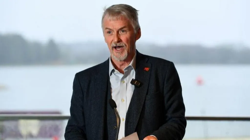 Huw stands on a podium with a body of water behind him. His mouth is open with a word and he is holding a paper in both hands He has a dark suit jacket and light blue shirt. He as short grey hair 