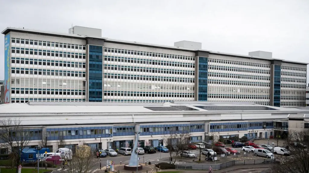 The outside of a large hospital, which is tall, blue and white with many windows, and car parked in front of it.