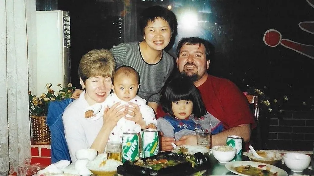 Three adults and two children pose for a photo. The adults are a white woman with short fair hair, a white man with dark hair and a goatee beard, and a Chinese woman with short dark hair. The fair haired woman is holding a baby and the man is holding a young child with black hair cut into a bob.