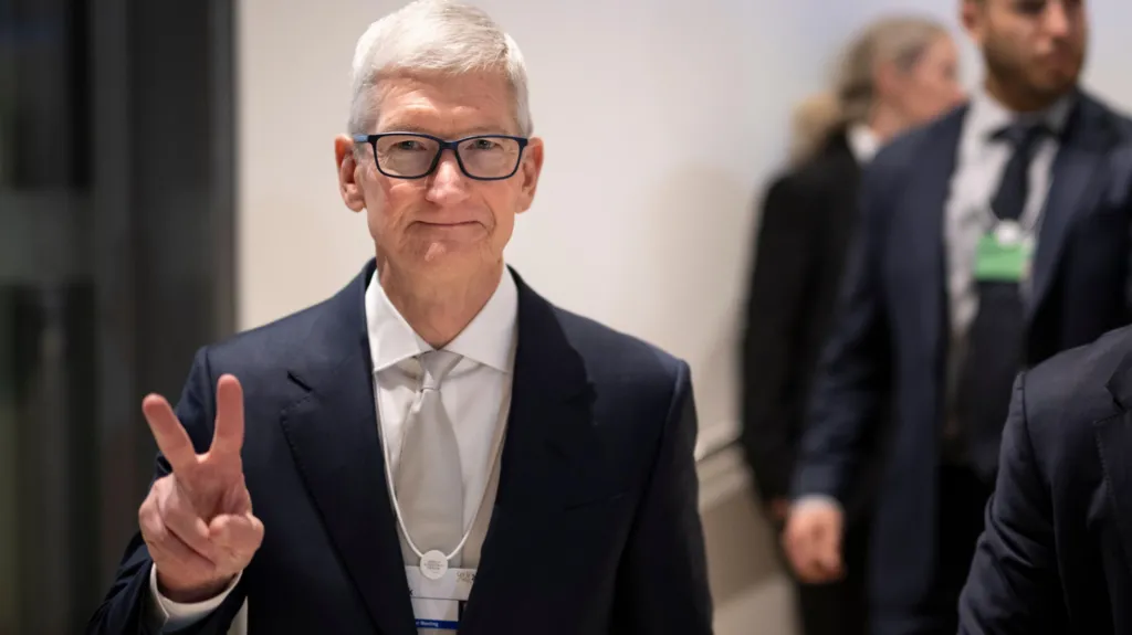 Tim Cook with short white hair, walking, flashing the peace sign wearing a black blazer and white collared shirt. 