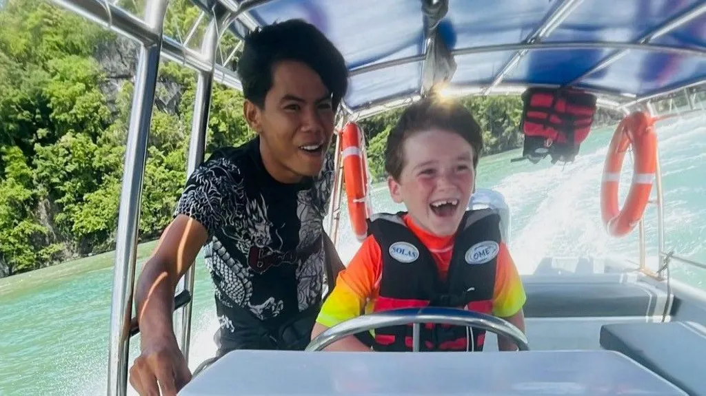 Alfie laughing while steering a boat. The sea and trees visible in the background