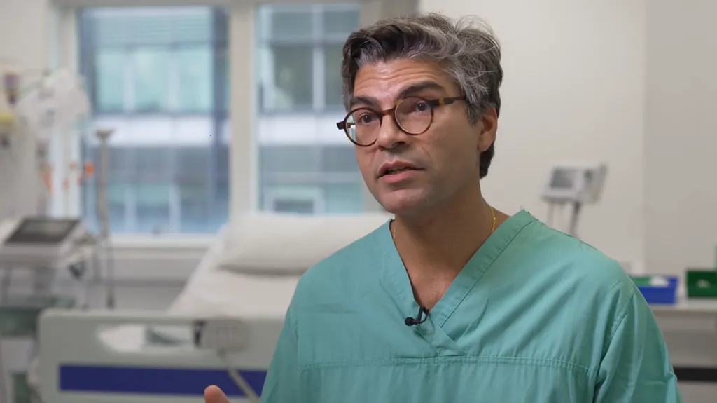 Ahmed Ahmed sat in a hospital room. Behind him is a hospital bed and clinical machines. He is wearing green scrubs and has on glasses. 