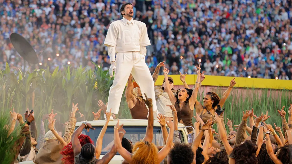 Bad Bunny on stage at the Superbowl. He is standing on a car and surrounded by female dancers waving their arms. 