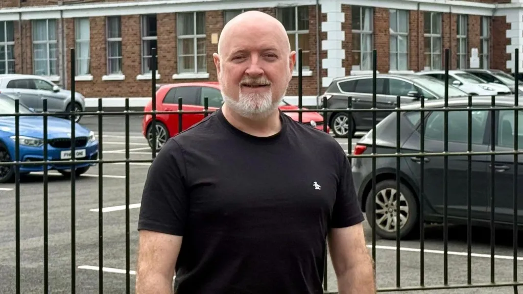 A bald man with a short white beard smiles close to the camera. He is wearing a black t-shirt with a small white logo on his left chest. Behind him is a black gate, with cars parked outside a red brick building with white corners.