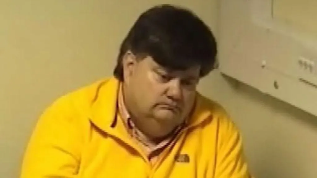 An old CCTV image of Carl Beech, with thick dark hair and a bright yellow fleece on, sitting in a room at a police station