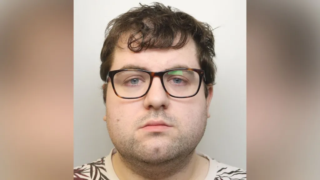 A custody image of Nathan Bennett. He has brown glasses on, a neutral expression, and mid-length brown hair. He is wearing a cream T-shirt with a brown, leaf-life design on it.