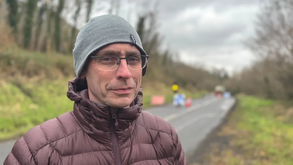 Paschal O'Shea stands by the roadside. He is warmly dressed in a brown puffy coat with a grey beanie hat. Behind him roadworks take place on the rural road. Traffic cones and temporary traffic lights can be seen in the distance. 