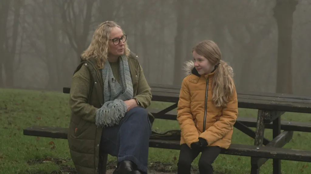 A picture of Cheryl Blenkinsop (left), with blonde hair, glasses and a green coat, sitting on a picnic bench next to her daughter Eliza (right), who has long blonde hair and is wearing an orange coat. There are trees in the background.