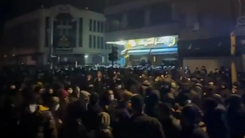 Screenshot from phone footage showing large crowd of people gathered outside after nightfall, with light from a building illuminating the dark
