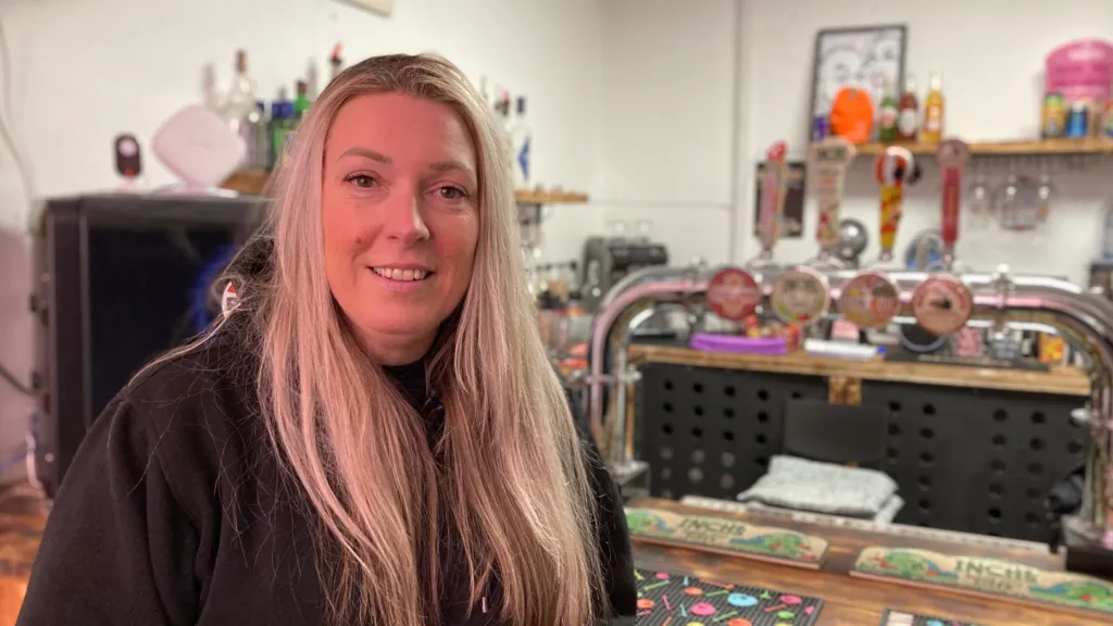Heather Dempsey, director of the Shadowbridge Brewery, standing at a bar. She has long blonde hair and is smiling at the camera. There are colourful beer mats on the bar, along with a number of draught beer pumps.