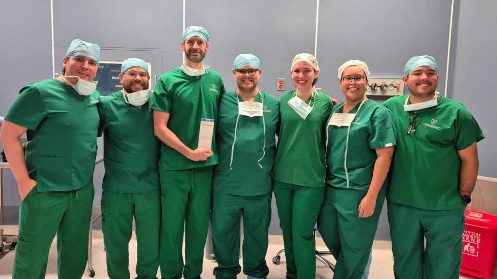 Seven surgeons stood looking at the camera for a photo wearing green scrubs and protective equipment. 