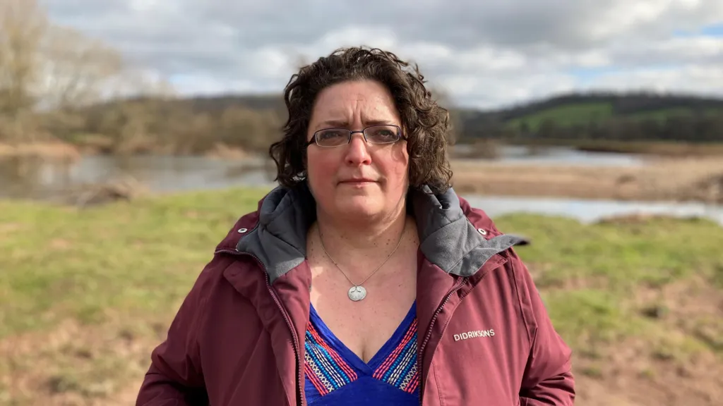 Gail Davies-Walsh  looks at the camera as she stands in front of a green bank and water body. she is wearing a burgundy windbreaker coat, open in the front revealing a purple v neck blouse. she has rectangular glasses and ear length curly dark hair