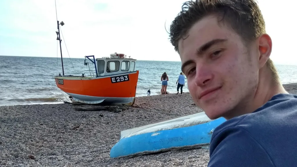 Gus sits on a beach in swimming shorts and his bare legs and a long sleeve t-shirt and looks over his left shoulder and smiles at the camera