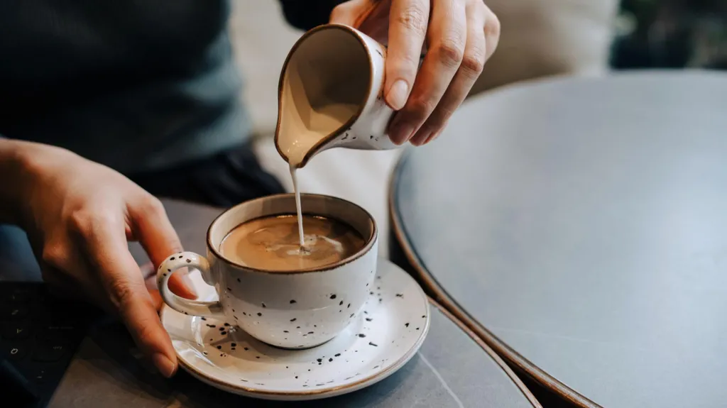 Milk from white jug with brown speckles on it being poured into a cup with black coffee in it. Cup is on a saucer and matches the jug. We can see two hands holding cup and jug. 