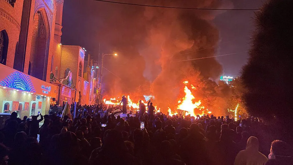 image of dozens of protesters gathered on a street in Tehran, Iran as cars burn in the background