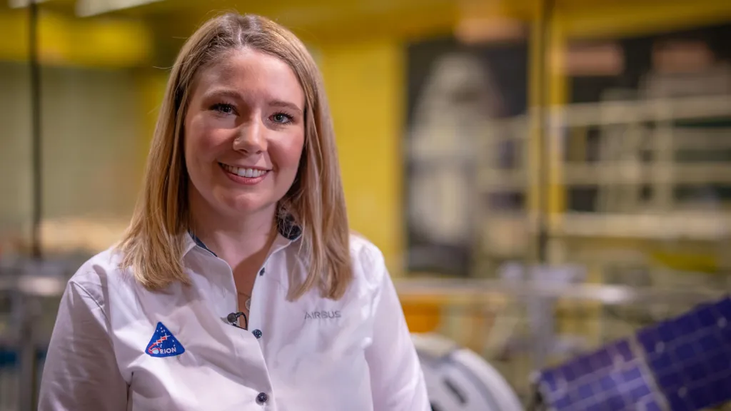 Airbus engineer Sian Cleaver standing in front of a cleanroom, which is blurred behind her in the background. She is wearing a white collared shirt with a small triangular Orion logo on it.