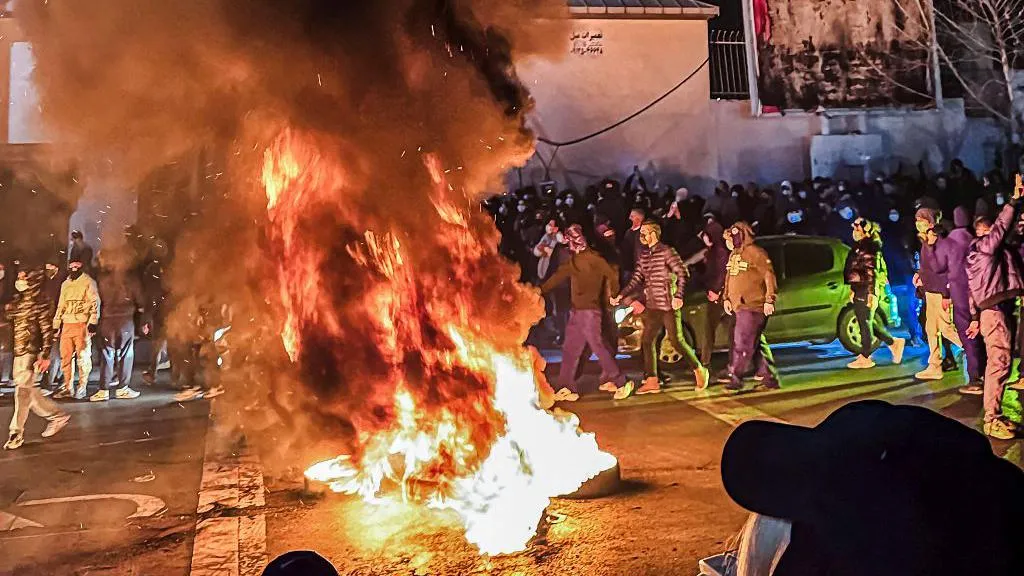 A large fire can be seen in the middle of a road, while cars are parked beside it. There are dozens of people surrounding the flames, some of them are wearing face masks, while others are not. 
