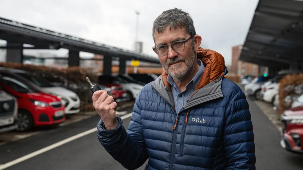 A man with dark hair, a light beard wearing a blue jacket, standing in front of parked cars with a car key in his hand