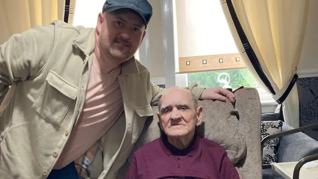 A man in a cap, Gary Mooney, poses next to a frail old man in a chair, William Mooney