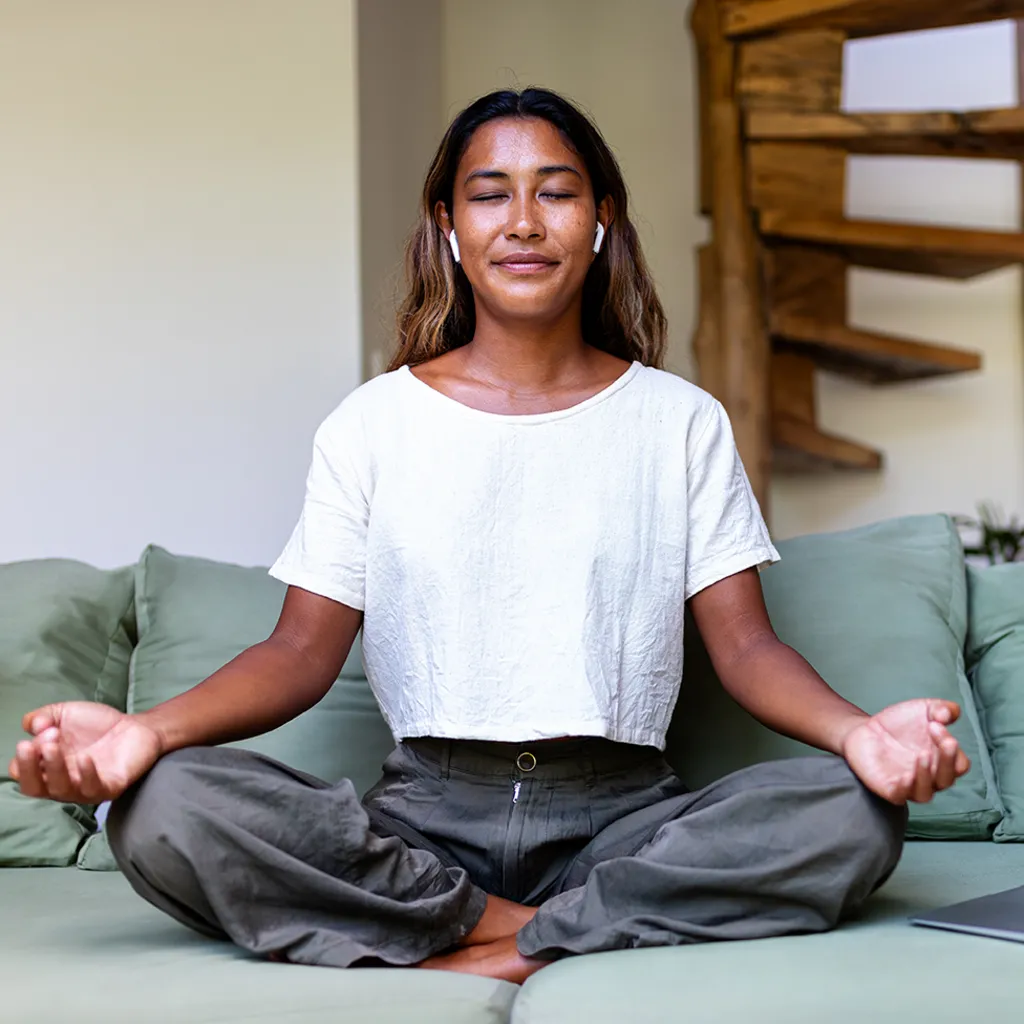 A woman sitting cross-legged on a green sofa. She is wearing a white T-shirt, grey trousers, and wireless earbuds. In the background, there is a wooden spiral staircase.