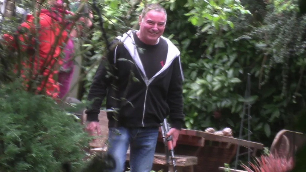 Thomas McKenna is smiling and is holding a replica AK47 assault rifle. He is wearing a black hoodie and blue jeans. There are bushes on either side. There are two people you can barely see behind him in brightly coloured clothing.