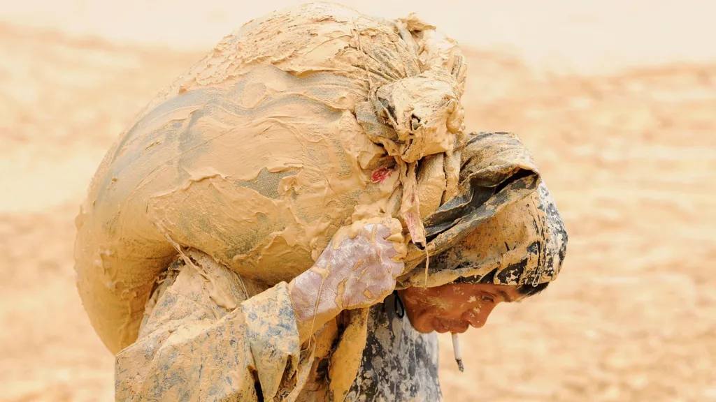 A man is bent over with a sack on this back. His clothes are covered and hands are covered in mud. In the background is a blurred dirt landscape.