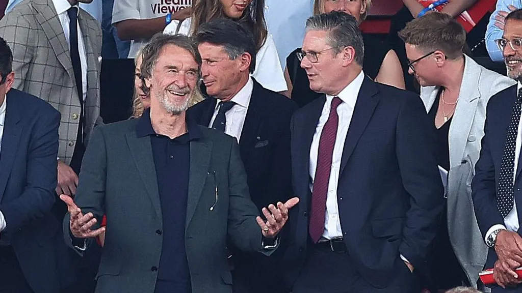 A group of suited men are seen in the Old Trafford football stands. Sir Jim Ratcliffe gestures with his hands in front of him, with Sir Keir Starmer next to him and Lord Coe behind them