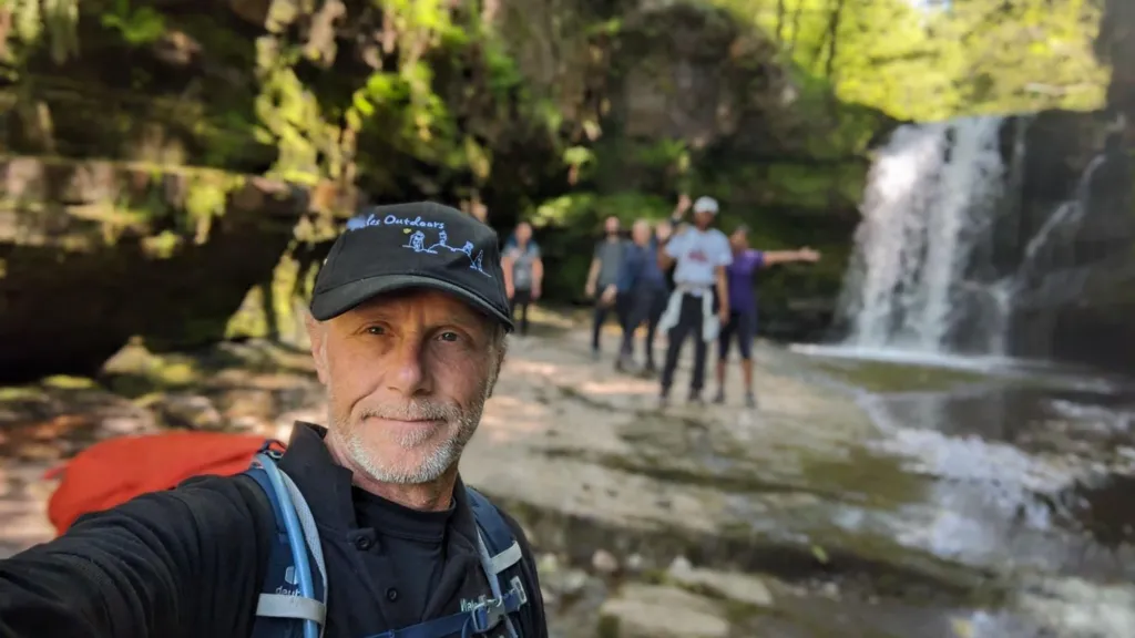 A man in the foreground wearing a cap and backpack is taking a selfie.  He is to the lower right section of the photo and behind him is a waterfall with a group waving.