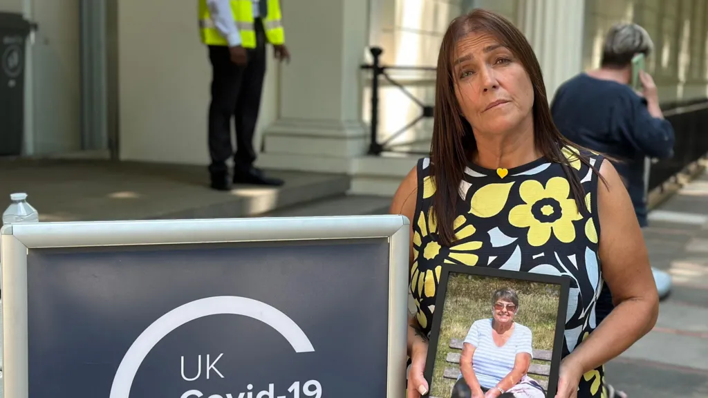 Nicky Hastie, in a floral summer dress, standing outside the Covid inquiry, holding an image of her mother Margaret.