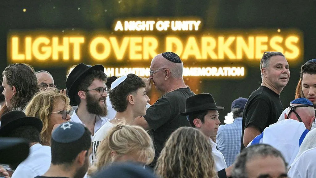 Mourners arrive to attend the memorial held for the victims of a shooting at Bondi Beach. A screen reading, "a night of unity", "light over darkness" can be seen in the background.