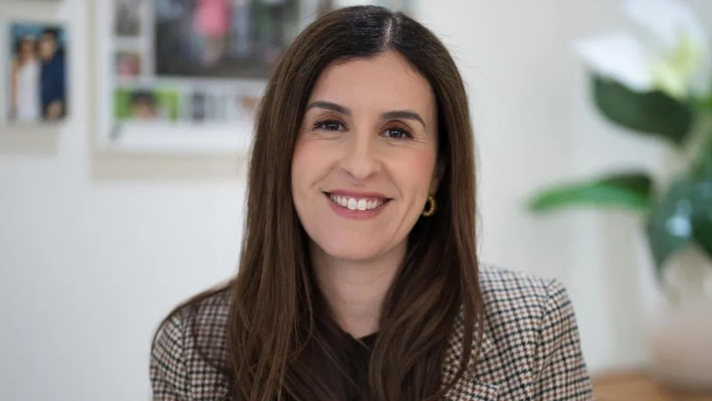 A woman with long dark hair and hoop earrings, wearing a check blazer, smiles at the camera.