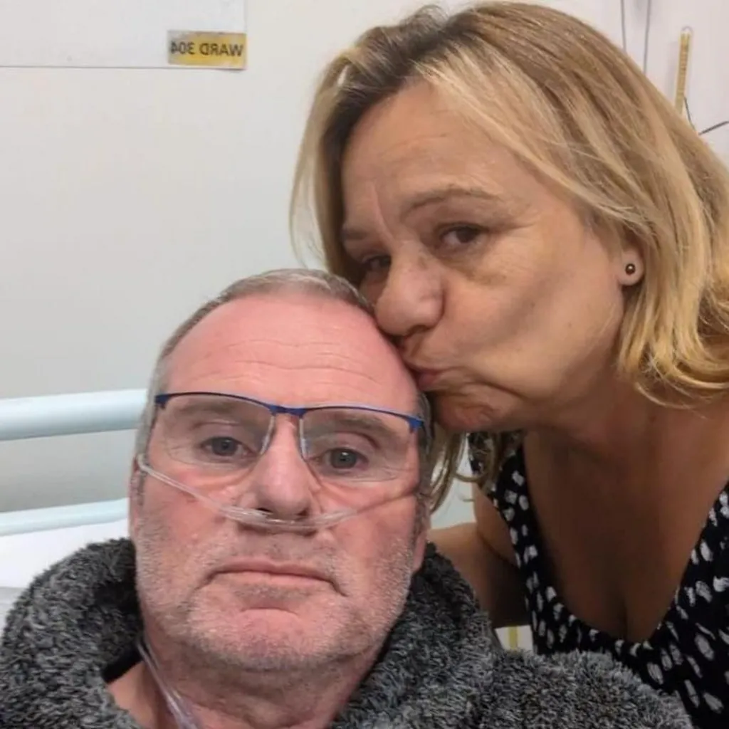 A man with a breathing tube in his nose looks at the camera while his wife, who has shoulder-length blonde hair, kisses him on the forehead. He is sat on a hospital bed.
