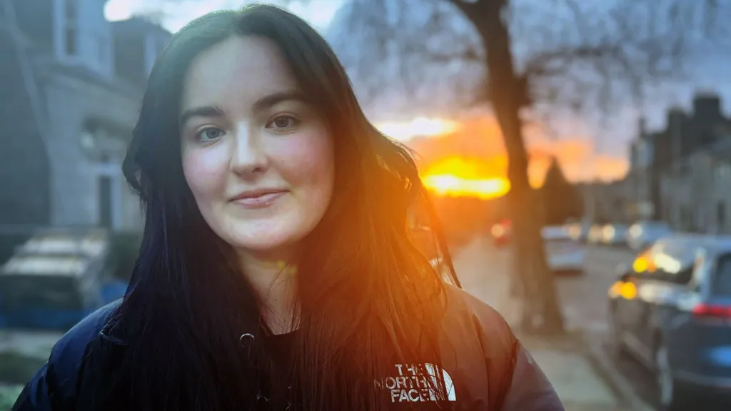Kate Finlay, young woman smiling at camera in street, with sun shining in the background.