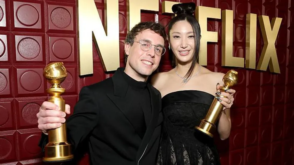 Mark Sonnenblick and Ejae pose for a photographer with their Golden Globe trophies. They are smiling and wearing black clothing in front of a maroon backdrop with Netflix written on it in large, gold letters.