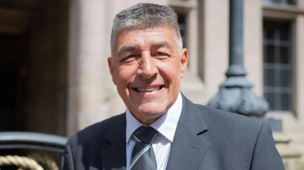 Chris Large is standing in front of the county council building, he is wearing a suit and tie and smiling at the camera.