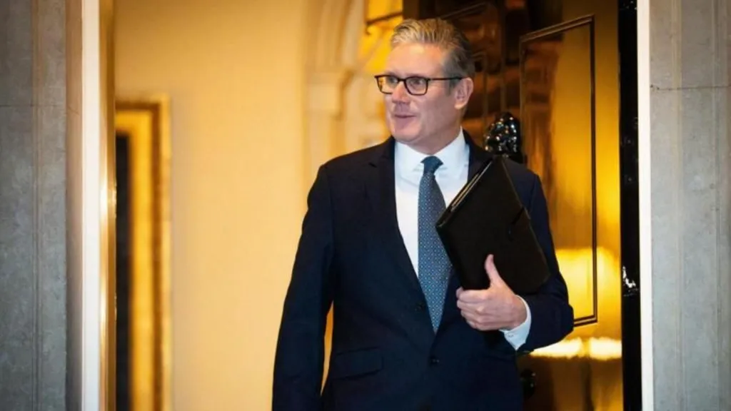 Keir Starmer walks out of the door to Number 10 Downing Street. He has short grey hair, glasses, and is wearing a navy suit and blue tie.