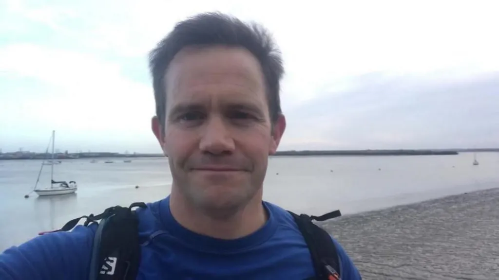 Mark Teeton is standing on a beach with water and a boat behind him. He is wearing a blue top and has a black rucksack. He is taking a selfie and smiling at the camera.