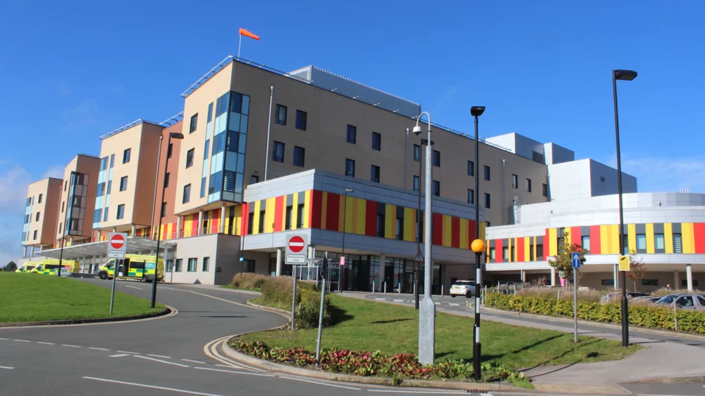A hospital with ambulances parked outside it. The hospital building is several storeys high, and has red and yellow cladding on the lower part.
